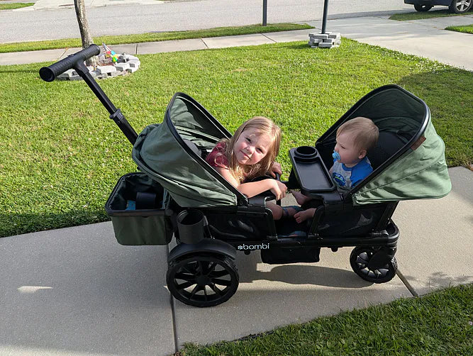 Two children sitting together in their green Waggle wagon on a sunny sidewalk