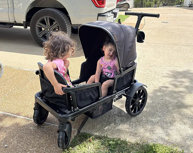 Two young girls riding in a black Waggle wagon beside a parked truck
